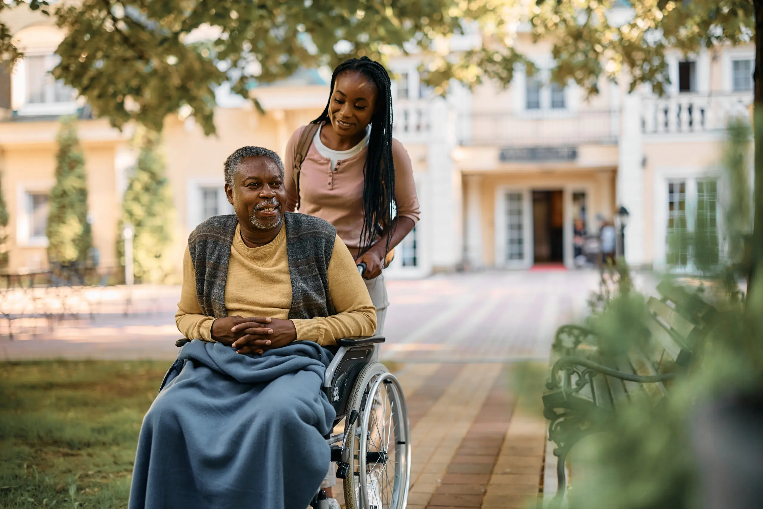 Happy African American senior man in wheelchair being pushed by his daughter through nursing home's park.
